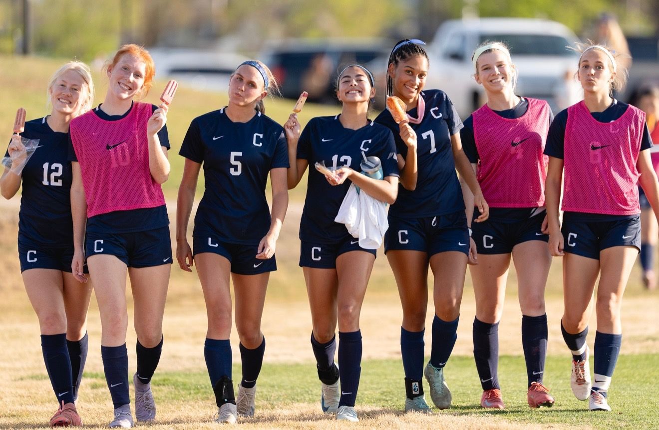 A group of seven female soccer players in blue uniforms and pink training vests walk on a field, smiling.
