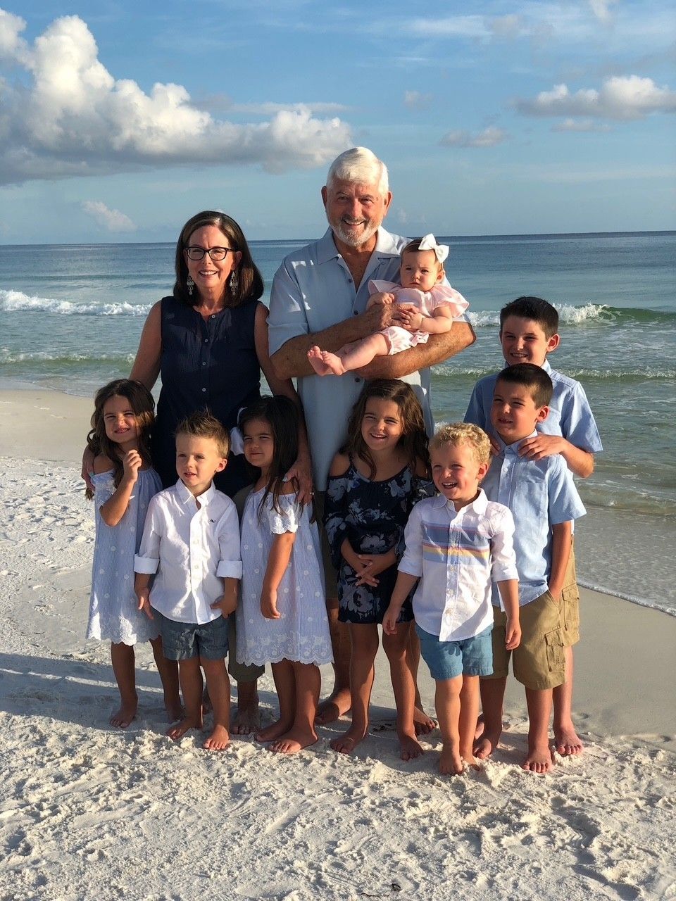 A man holds a baby on a beach, facing the ocean. The baby wears a pink dress, and the man has white hair and a blue shirt.