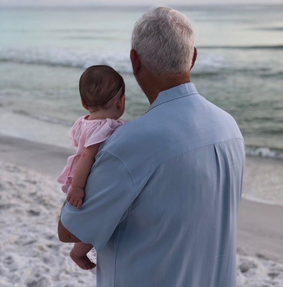 A man holds a baby on a beach, facing the ocean. The baby wears a pink dress, and the man has white hair and a blue shirt.