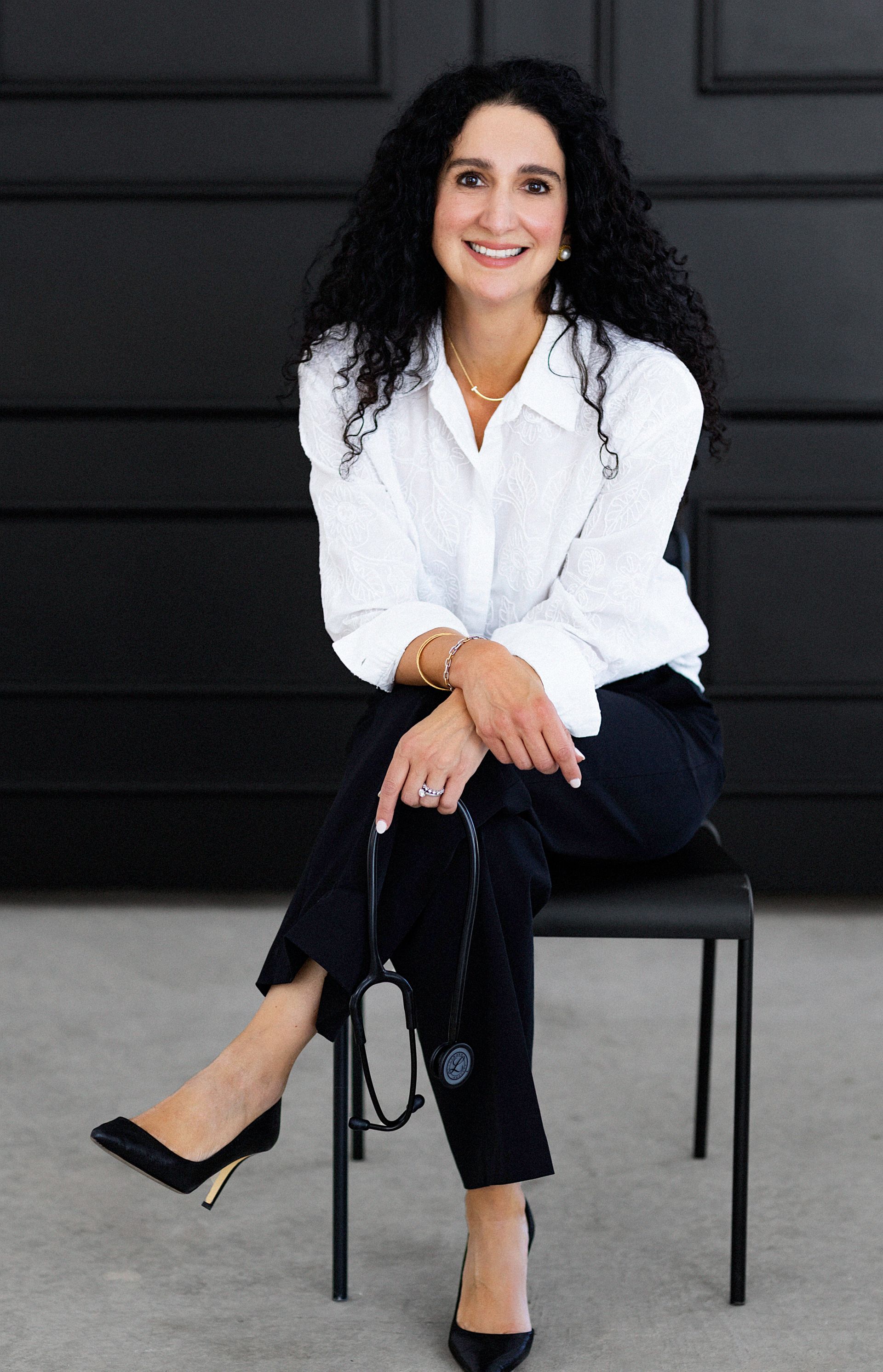 Woman with dark curly hair smiles, wearing a white blazer over a green top. She has a gold bracelet and a necklace.