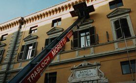 Vista esterna di un edificio; un ascensore solleva i mobili fino a una finestra; sull'ascensore è stampato 