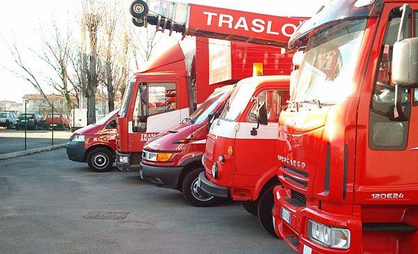 Camion rossi per le consegne parcheggiati in fila, il primo con un sollevatore, sotto un cielo luminoso.
