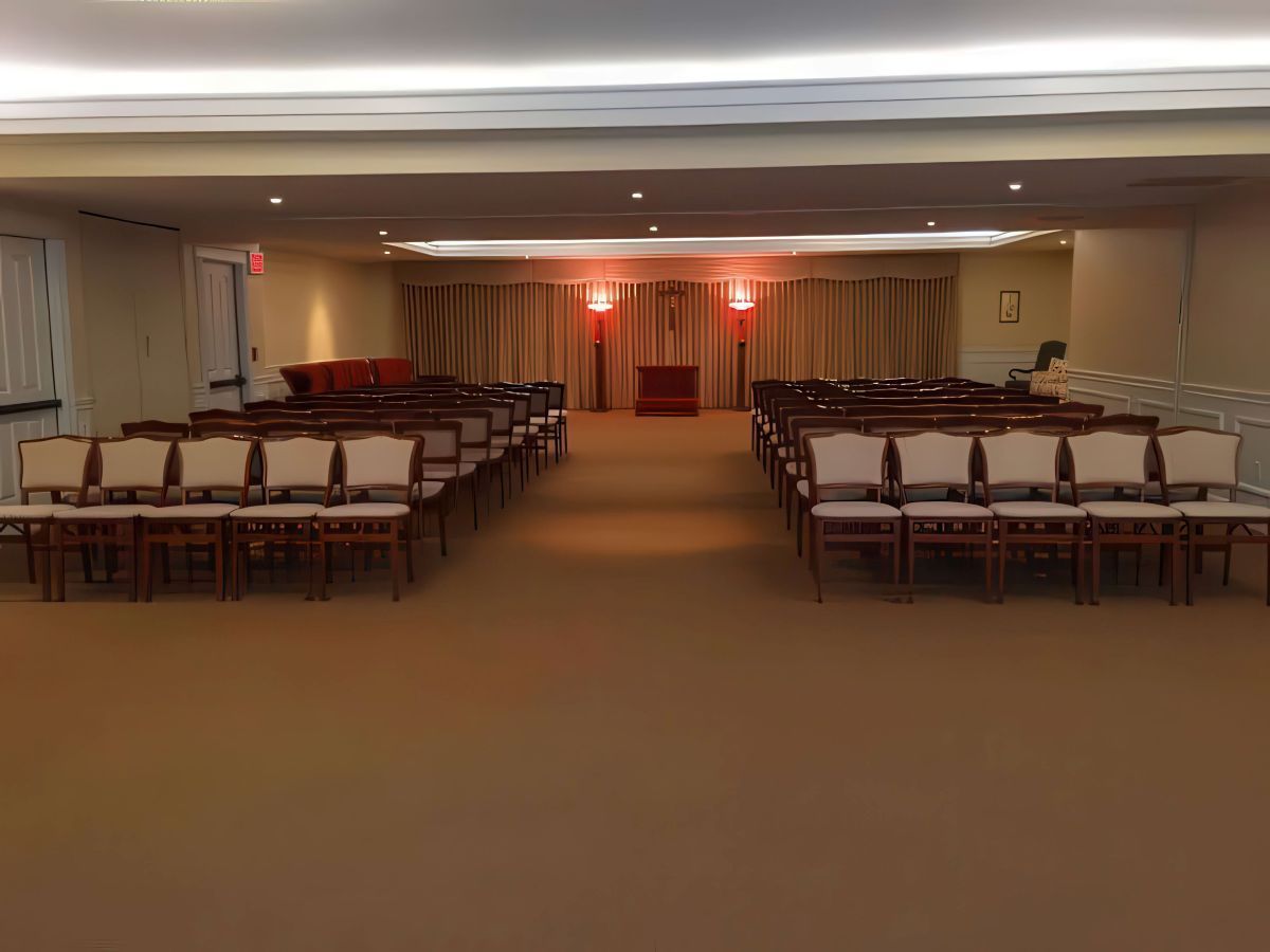 Interior of a beige room with rows of chairs facing a stage with a red cloth and curtains.