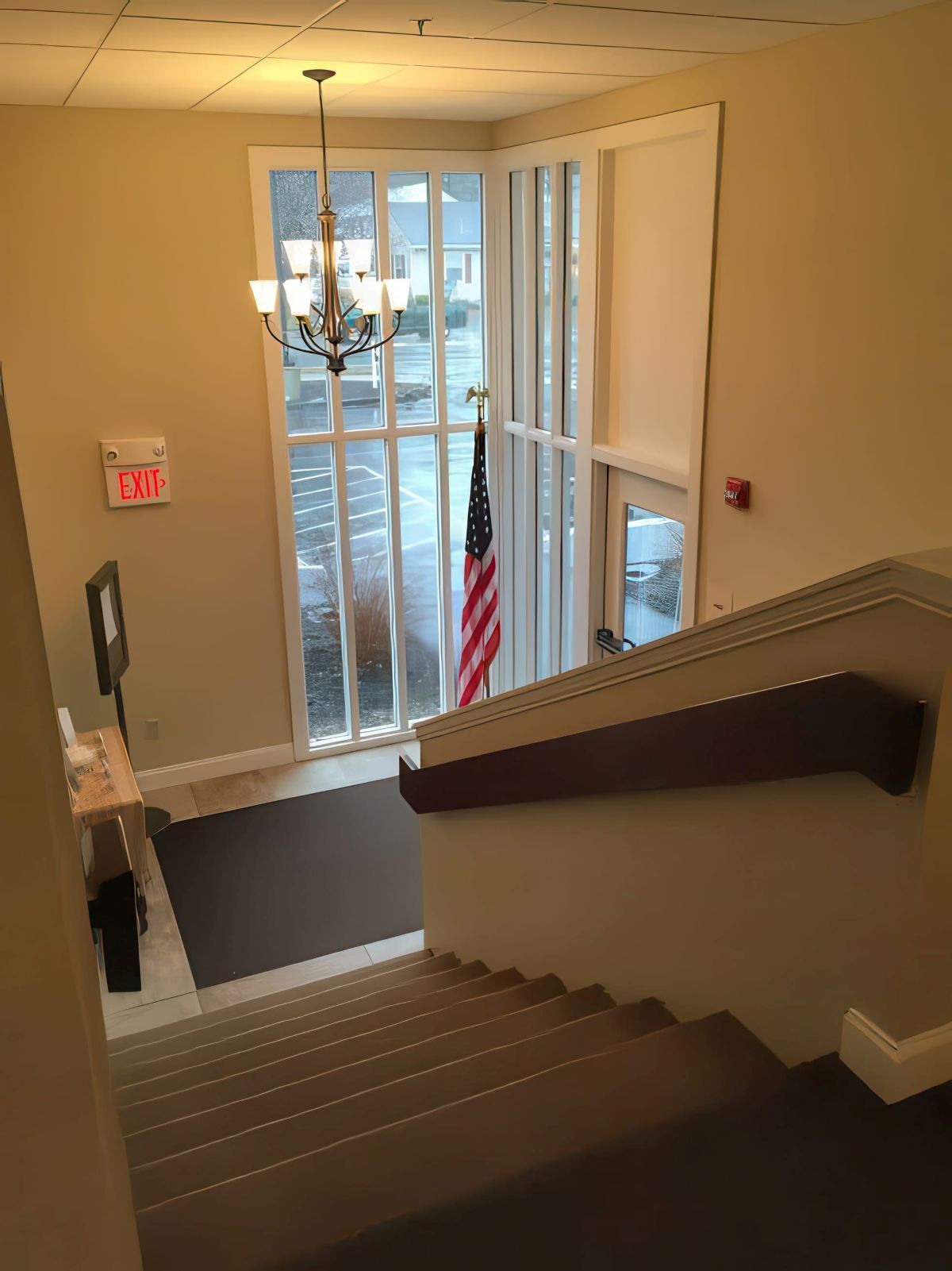 Stairwell leading down to a lobby with an American flag and large window. Tan walls, brown carpet, chandelier.