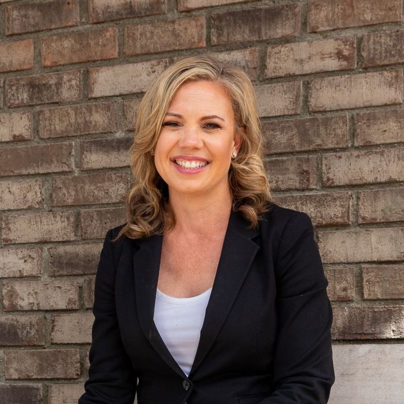 A woman in a black jacket is smiling in front of a brick wall.