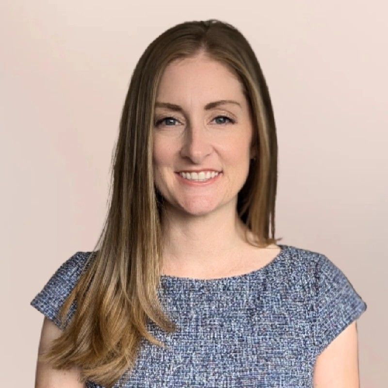 Woman with long blonde hair, wearing a blue top, smiling at the camera. Light pink background.
