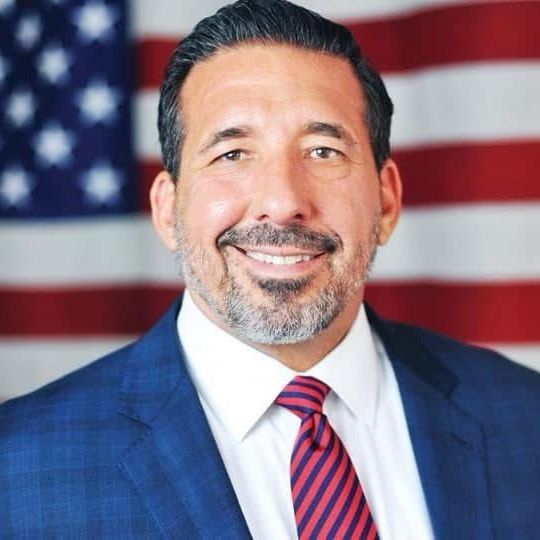 A man in a suit and tie is smiling in front of an american flag.