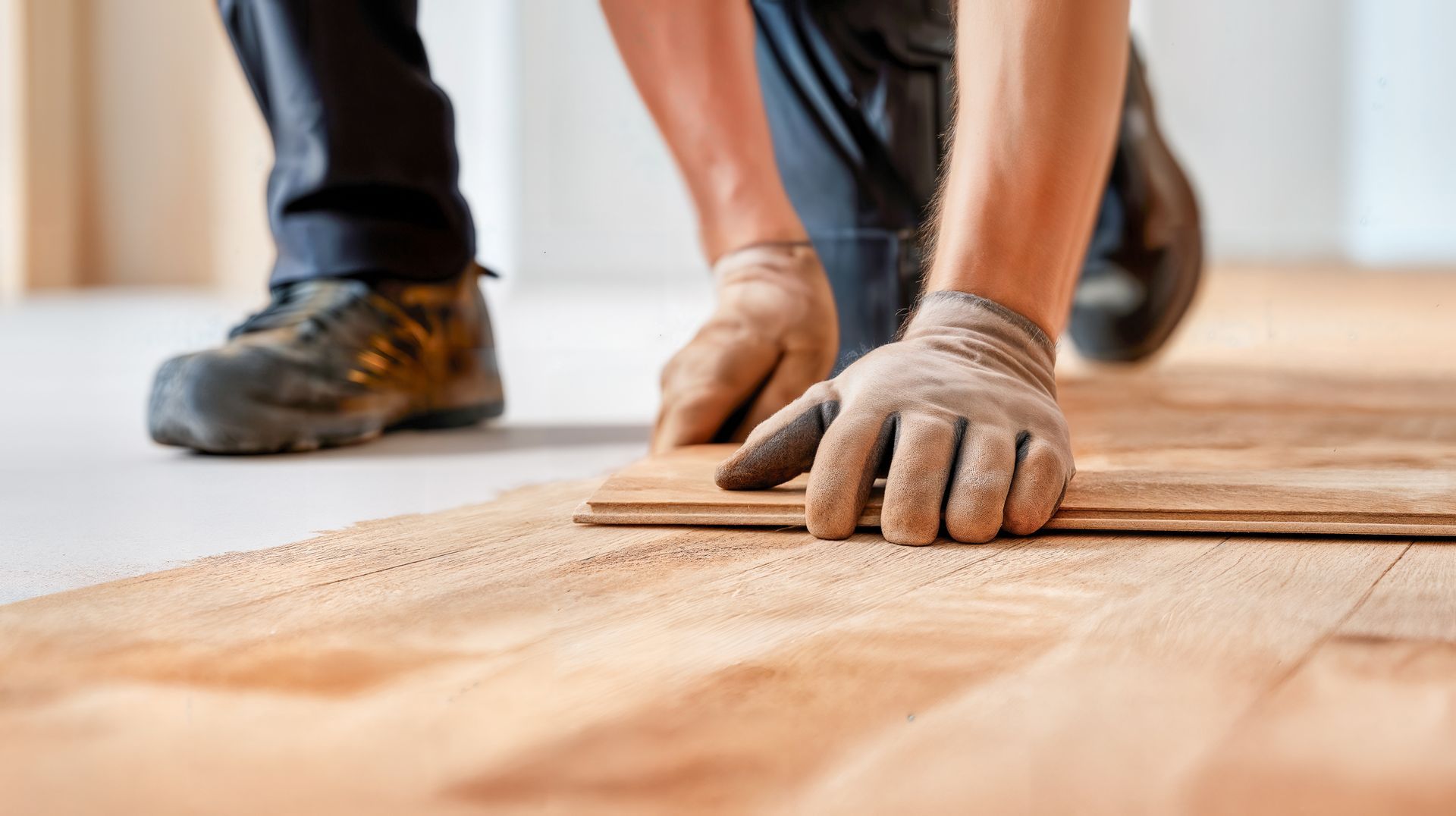 A worker kneeling on a floor installs light-colored wooden planks wearing work gloves.