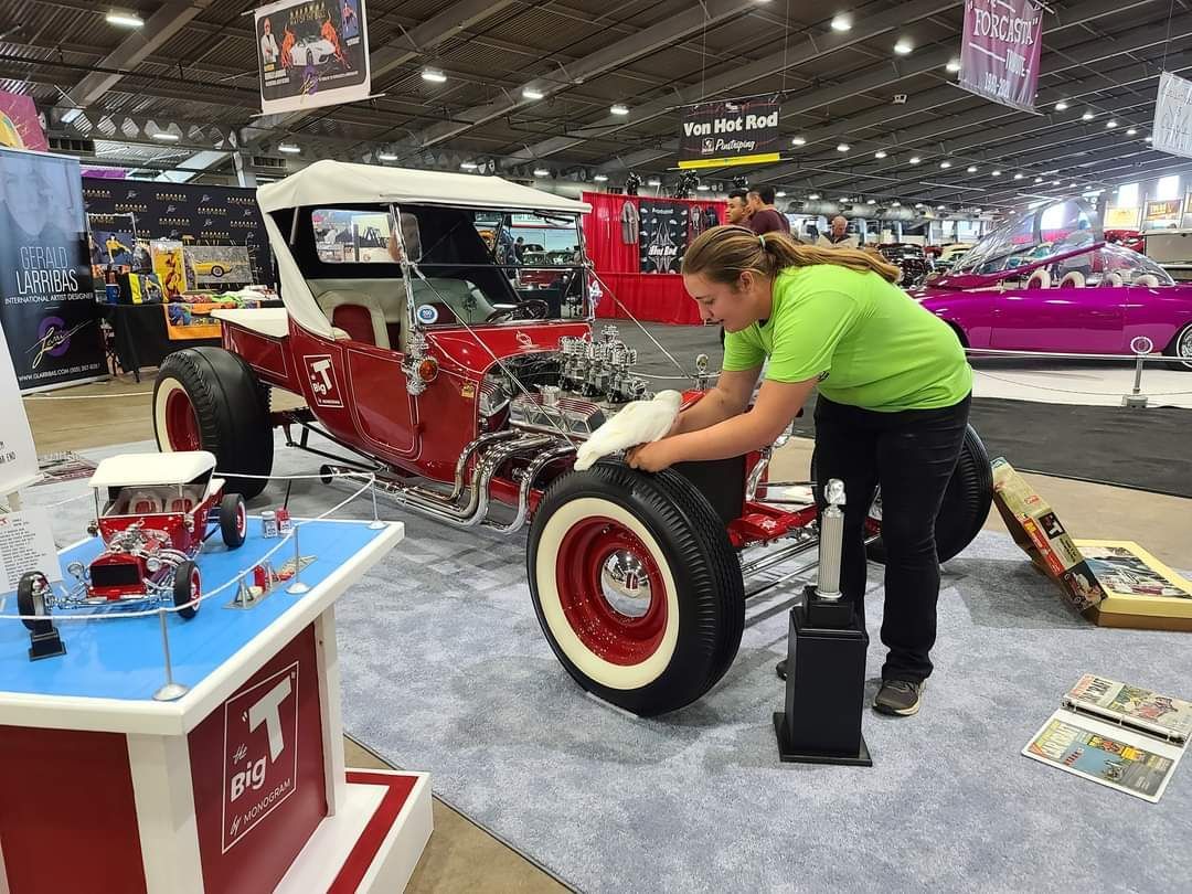 Woman cleaning a red and white hot rod at an indoor car show.