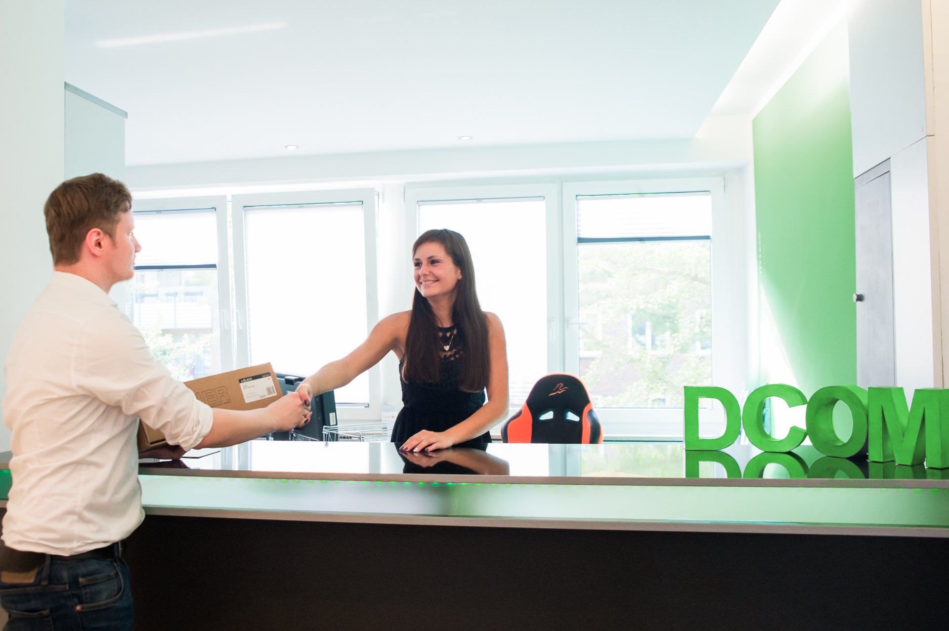 Ein Mann übergibt einer Frau am Empfang ein Paket. Grüne Akzente, neutrale Farben. Lächelnde Gesichter, Büroumgebung.