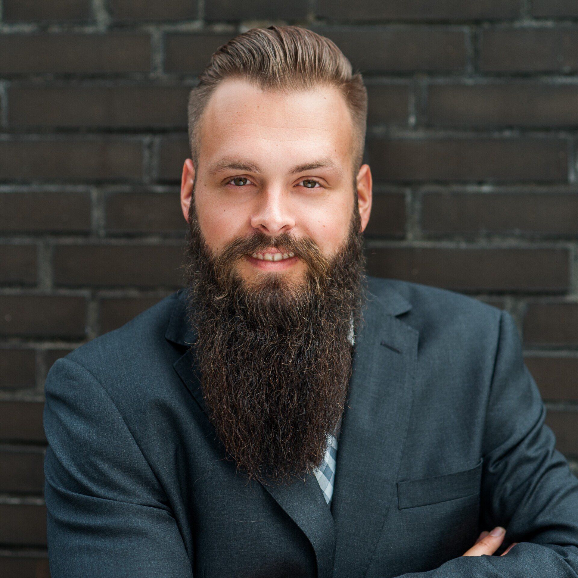 Ein Mann mit Vollbart im Anzug lächelt mit verschränkten Armen und lehnt an einer dunklen Backsteinmauer.