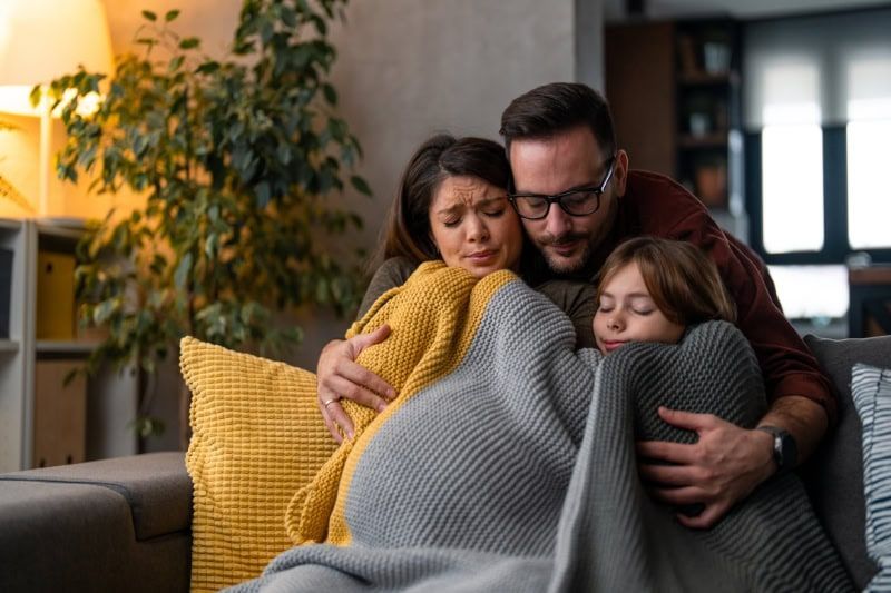 A family is sitting on a couch wrapped in a blanket.