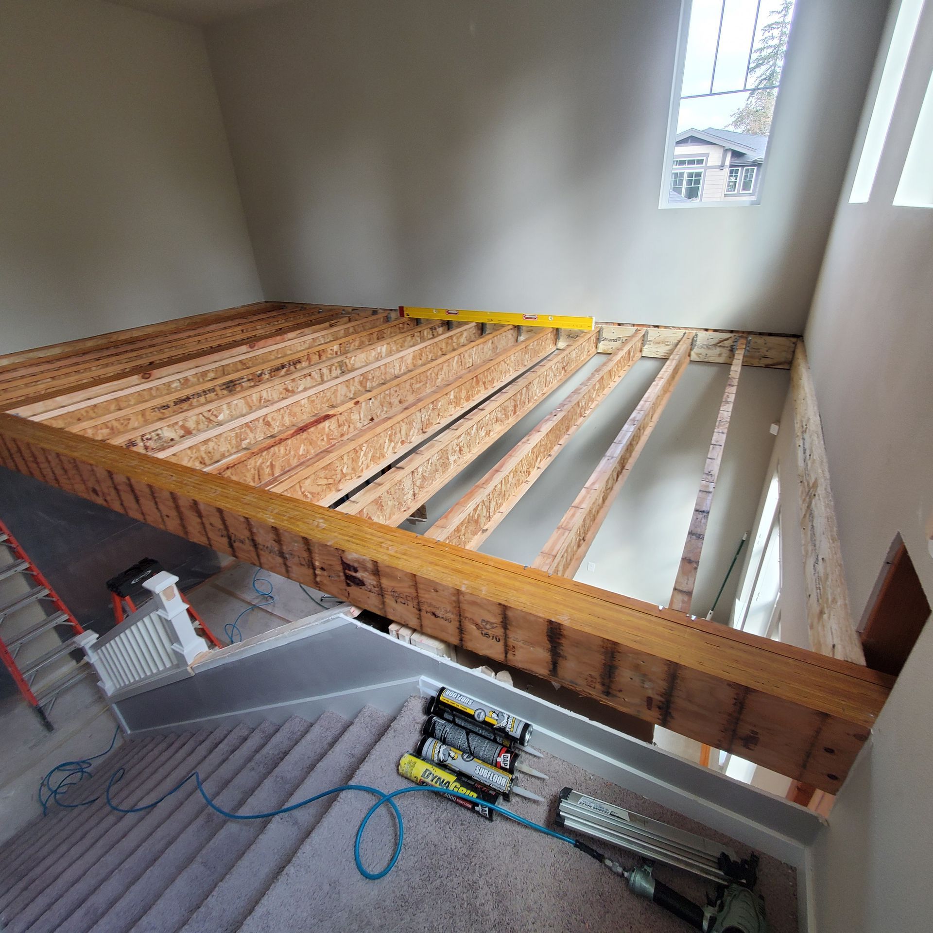 Construction of a wooden loft over a stairwell, unfinished. Light-colored walls and carpet, nail guns and tools.