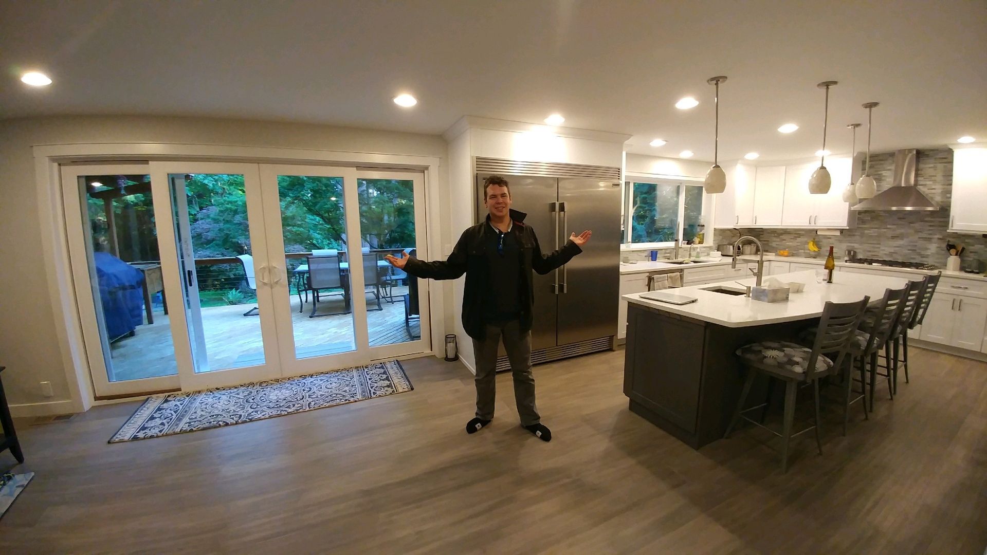 Man standing in a modern kitchen, arms outstretched, next to large sliding doors and an island with stools.