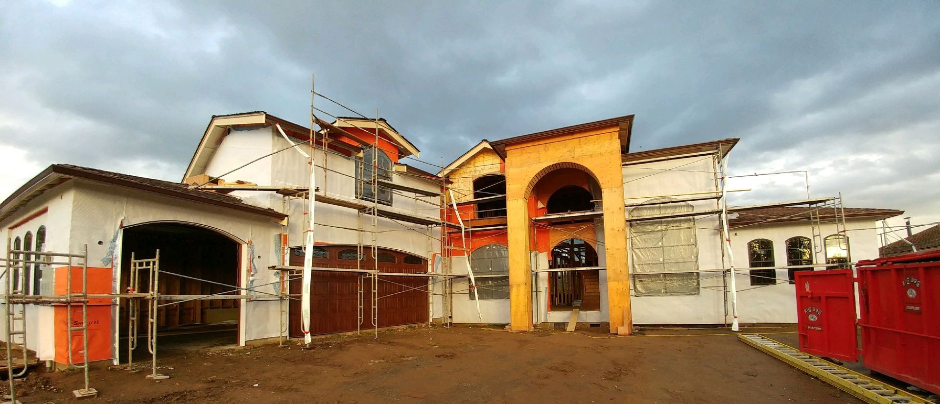 A house under construction with orange accents, brown garage doors, and scaffolding against a cloudy sky.