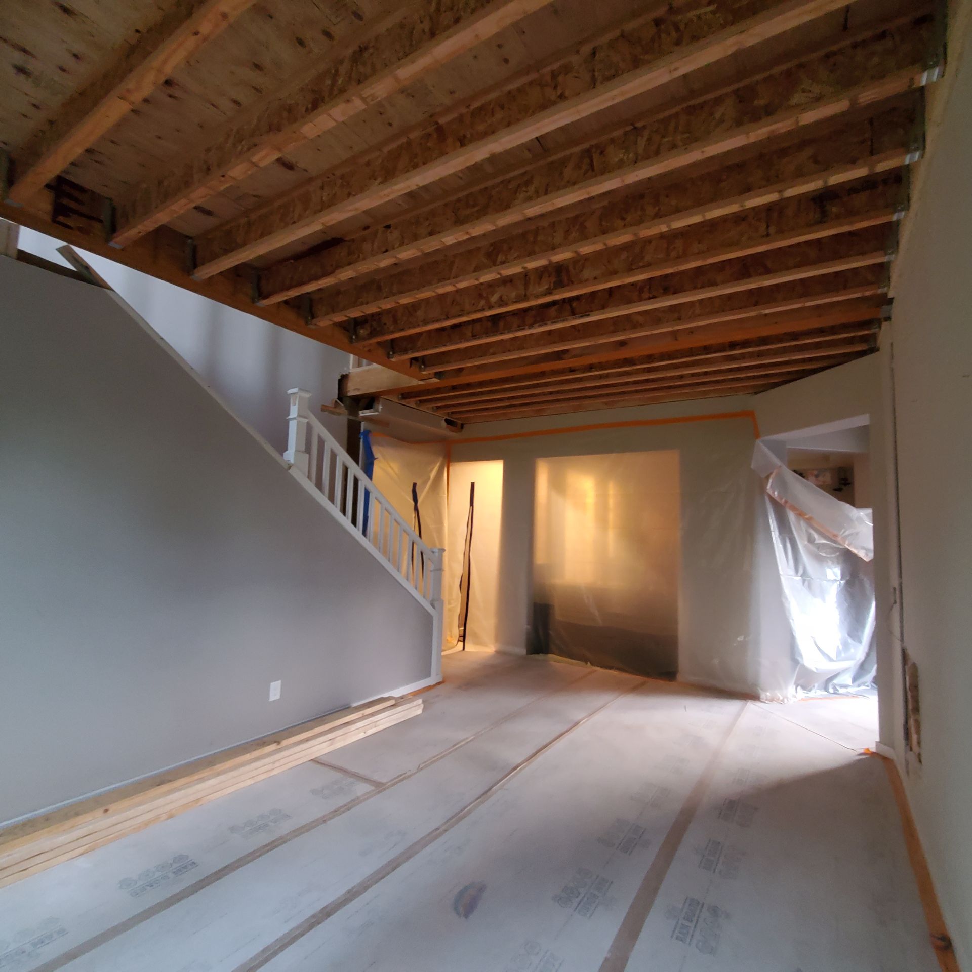 Interior under construction: unfinished staircase, exposed ceiling beams, plastic sheeting, light gray walls