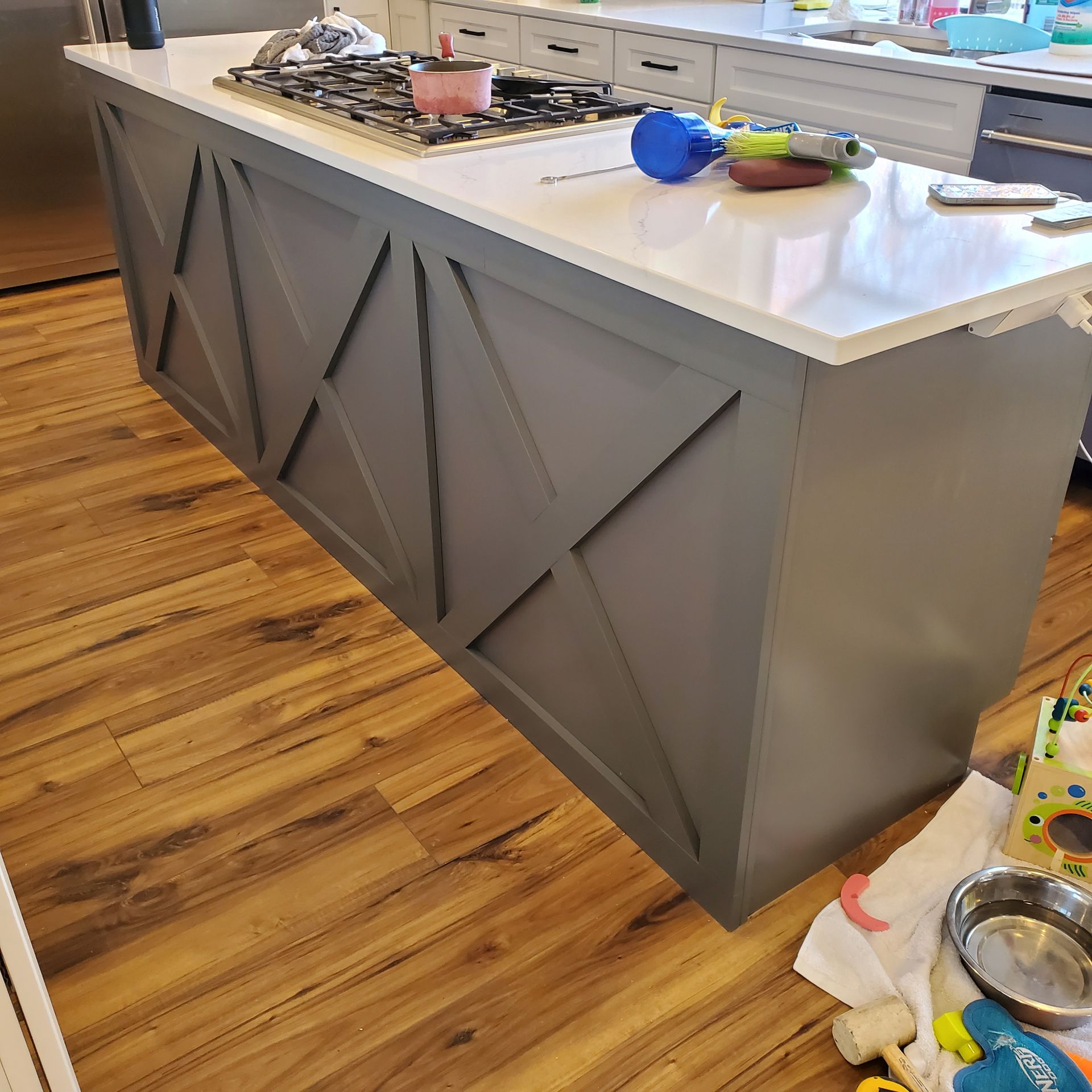 Kitchen island with gray X-paneling, white countertop, and a stovetop. Brown wood floor.