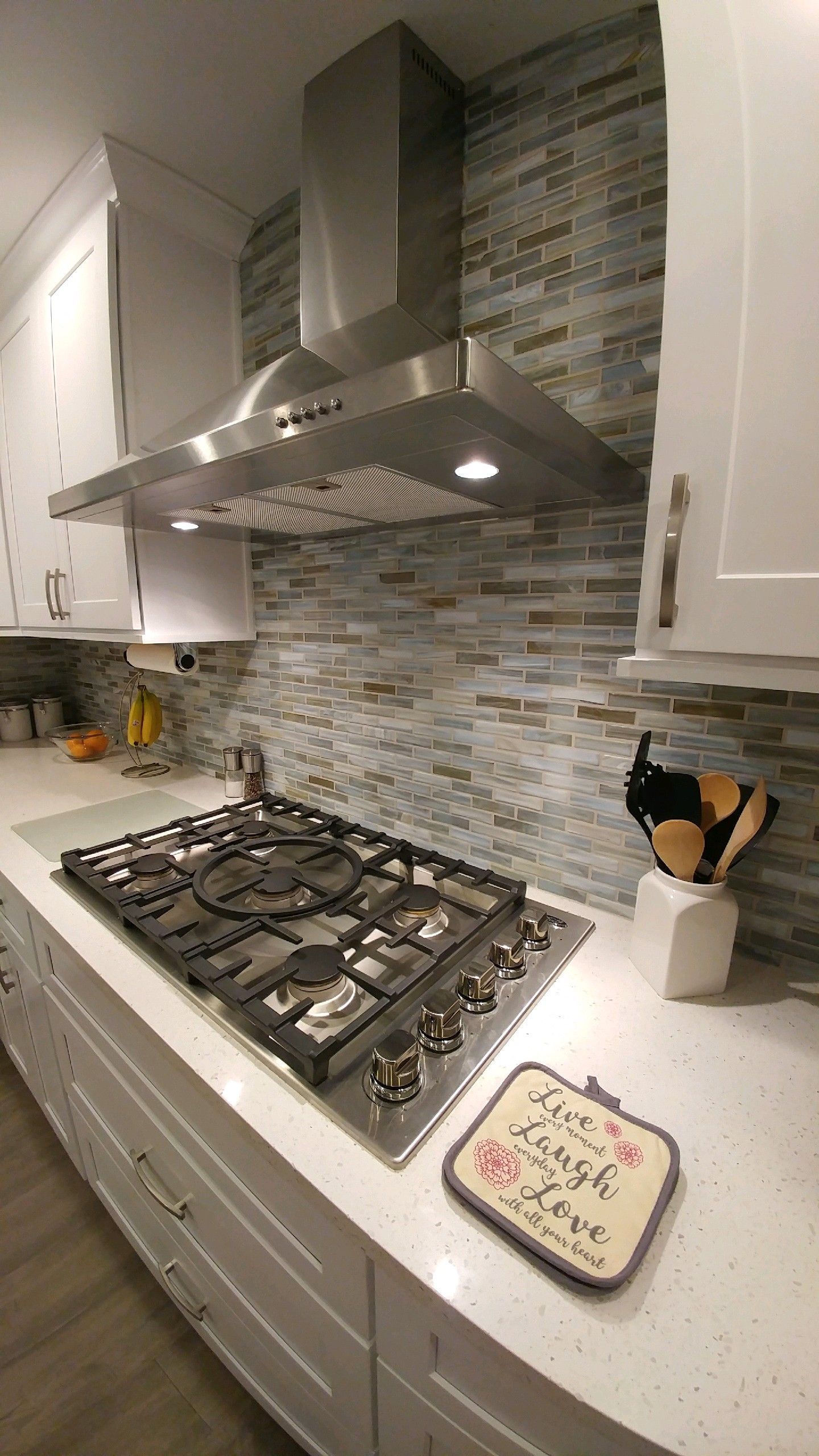 Stainless steel range hood over a cooktop in a white kitchen with glass tile backsplash.