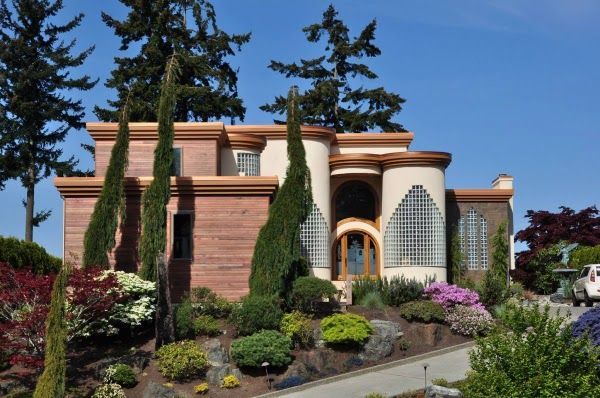 House with beige and red facade, arched windows, and lush landscaping under a blue sky.