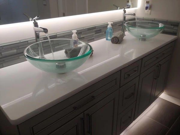 Bathroom vanity with two glass bowl sinks; faucets running, white countertop, gray cabinets, tile backsplash.