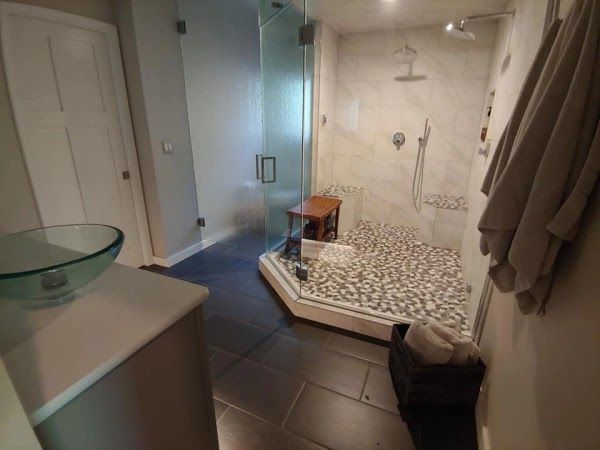Modern bathroom with glass shower, pebble floor, dark tile, light gray walls, vanity, and towels.