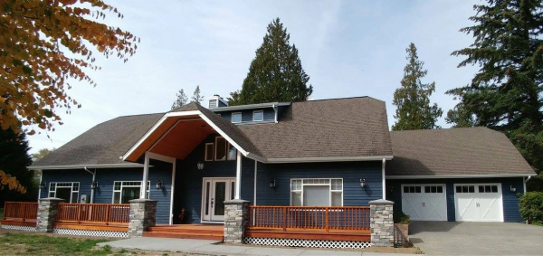 Blue house with brown roof and wooden porch. Two-car garage, trees in background.
