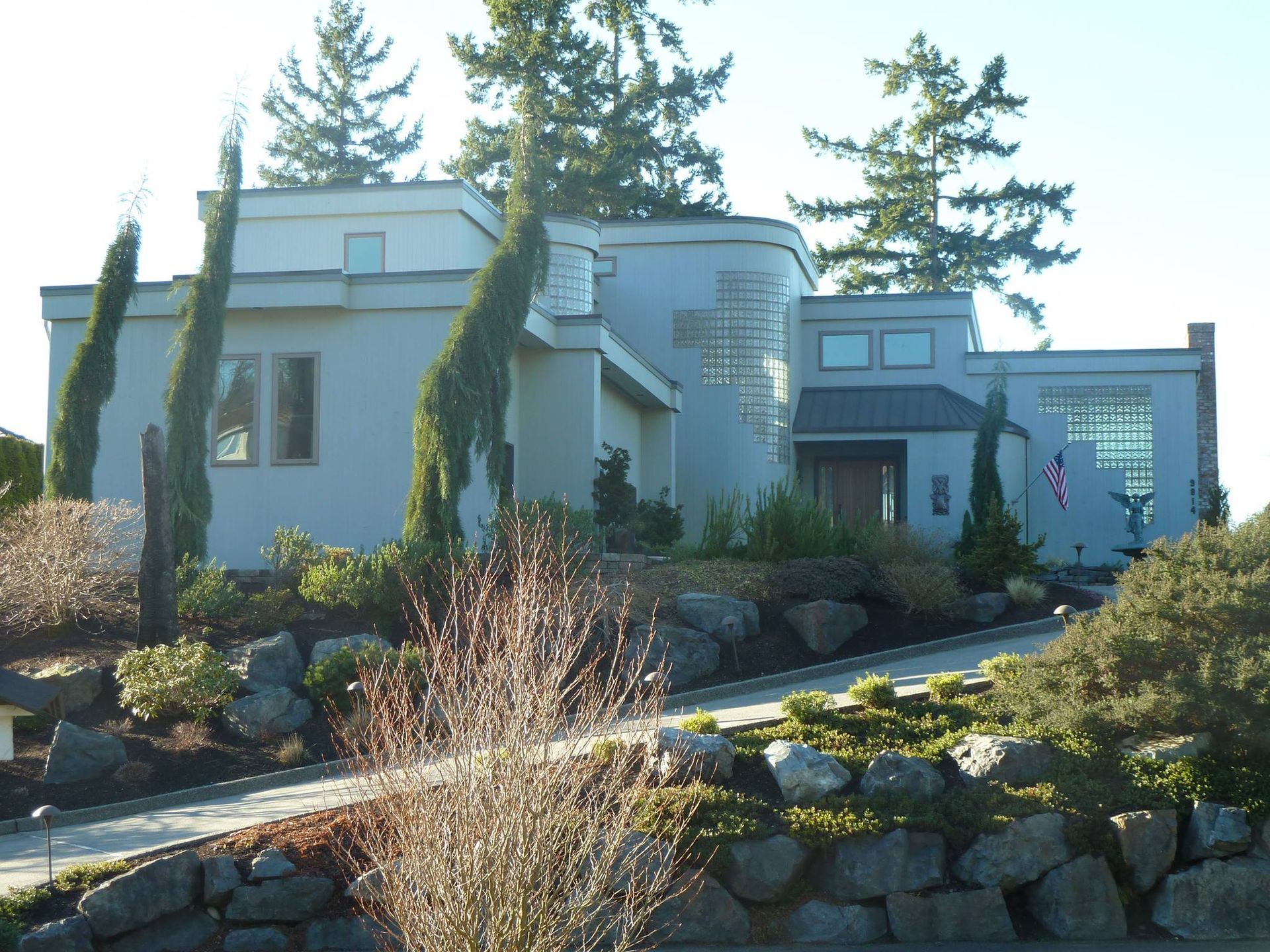 Modern gray house with stone retaining wall, set amongst lush greenery.