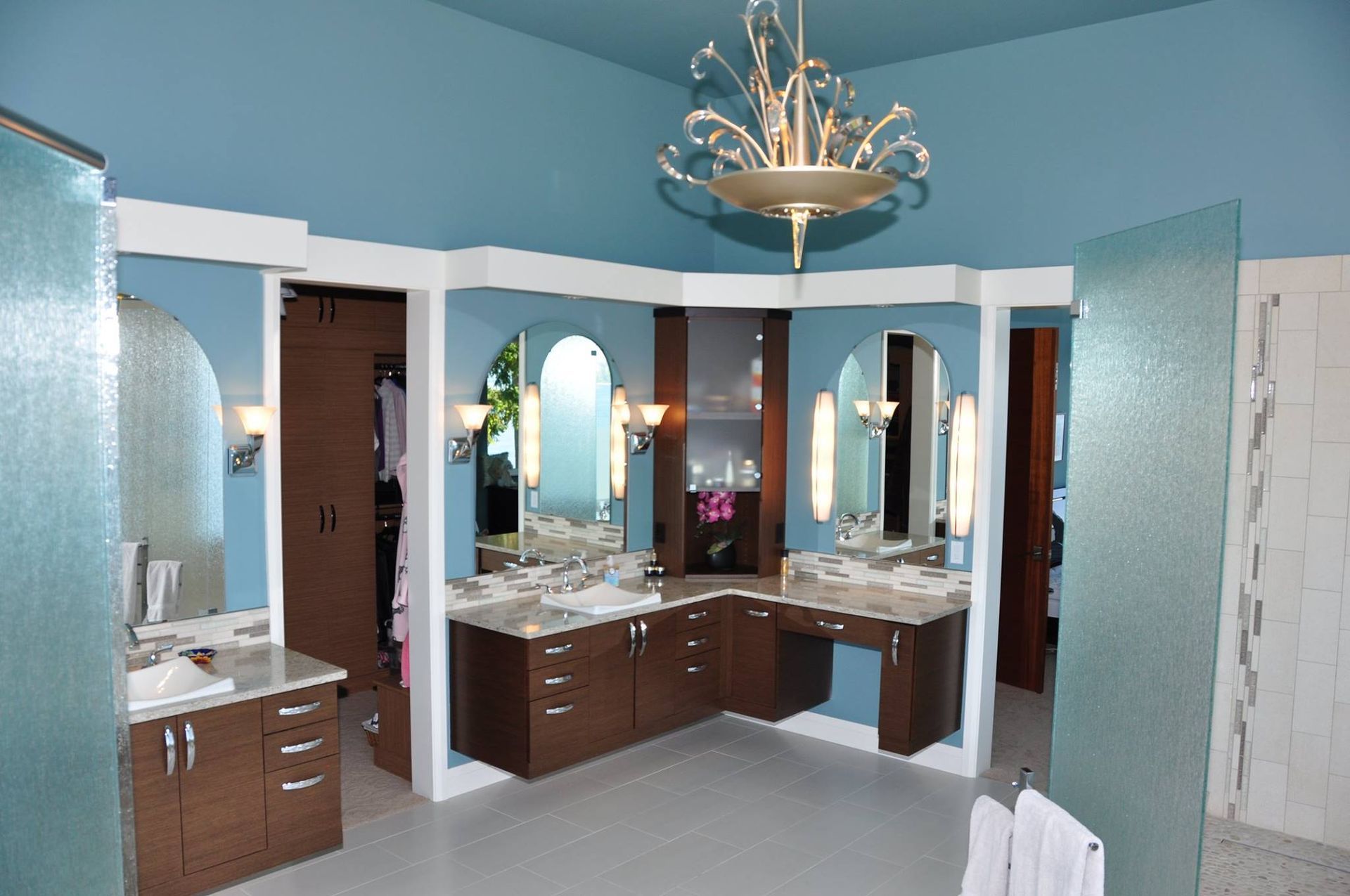 Large bathroom with blue walls, brown cabinets, and glass shower walls; two sinks, and a chandelier.