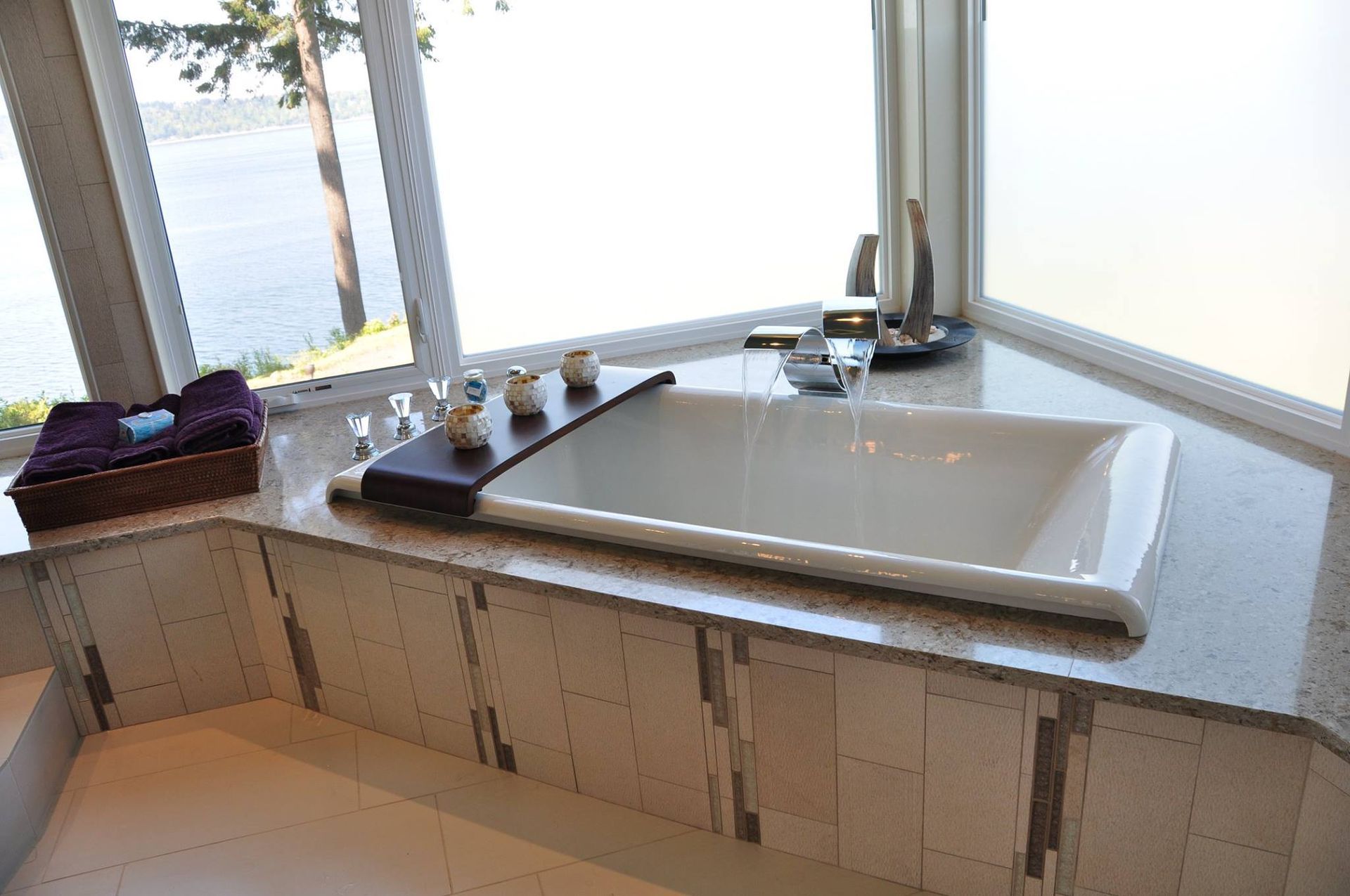 Corner bathtub with lake view, white tub, dark wood tray, stone counter and trim.