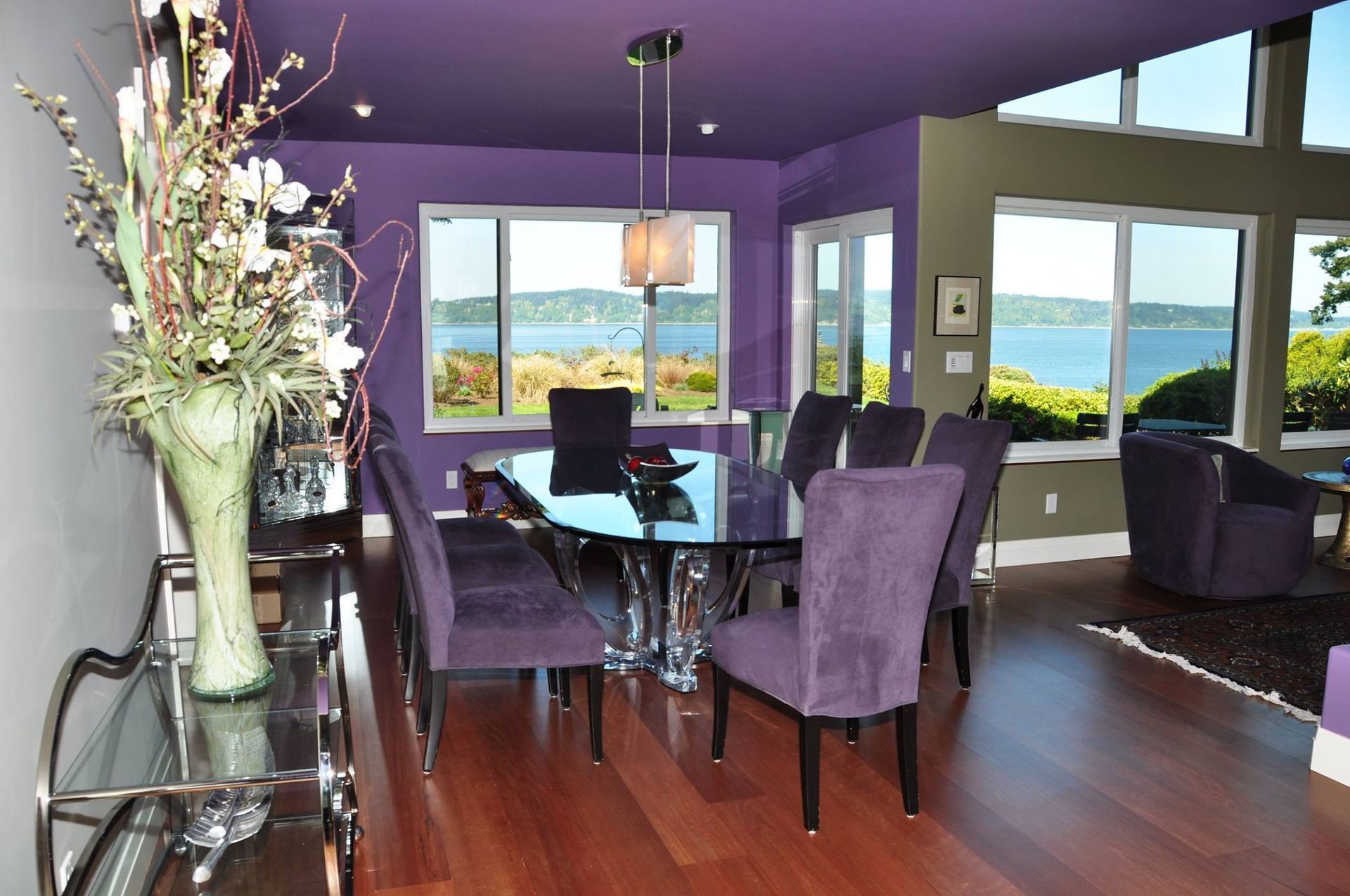 Purple dining room with glass table, purple chairs, and a water view.