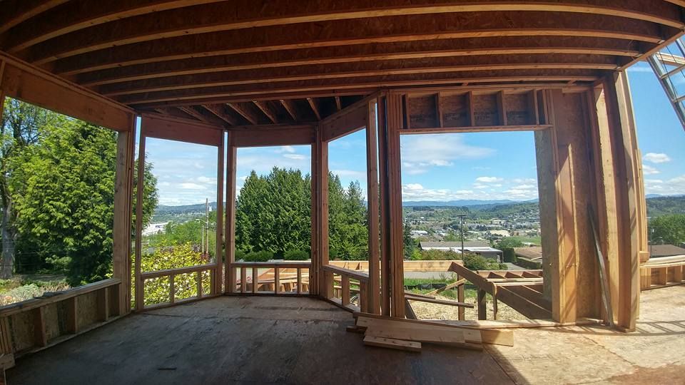Framing of a house under construction with large windows overlooking a city and trees on a sunny day.