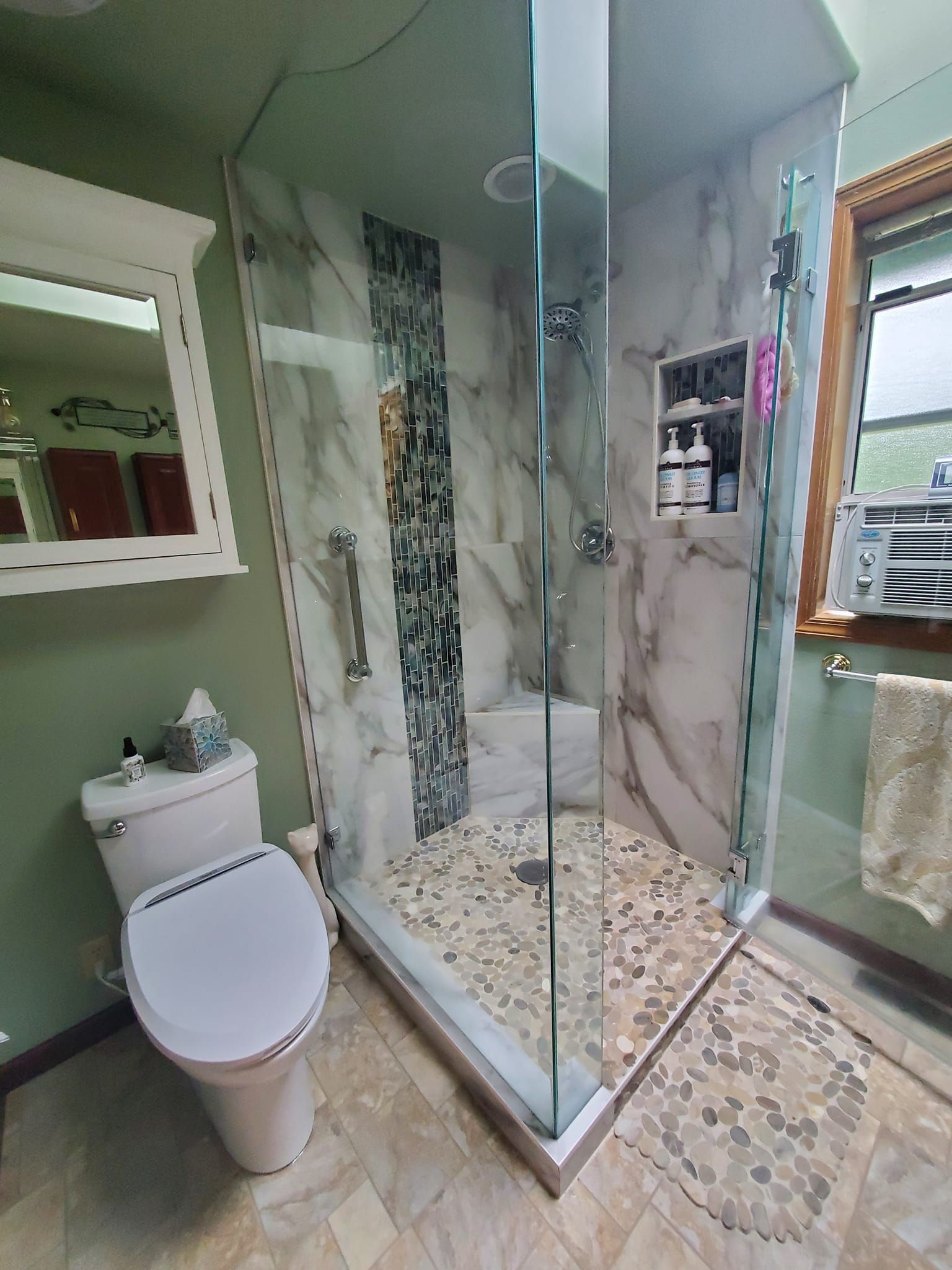 Bathroom with glass shower, marble tile, and pebble shower floor. Toilet and vanity are visible.