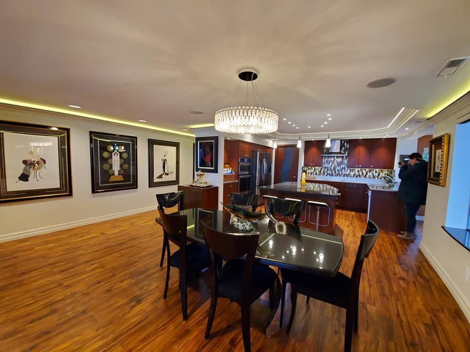 Dining room with dark table and chairs, art, and kitchen visible in the background. A person takes a photo.