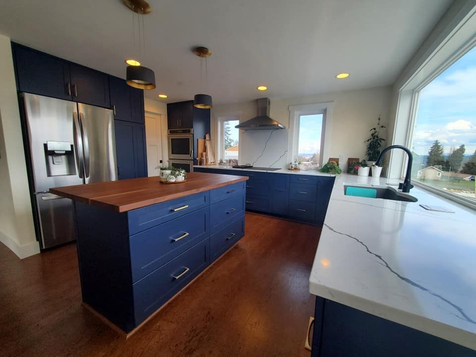 Blue kitchen with island and dark wood countertop. Stainless steel fridge, large window, and wood floors.