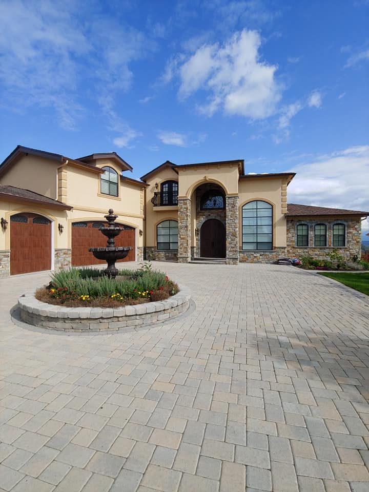 Beige stucco house with stone accents and a fountain in the paved driveway under a blue sky.