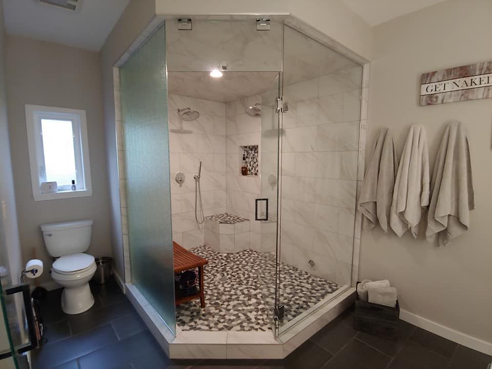 Spacious bathroom with large glass shower, pebble floor, and white marble walls. Gray walls, toilet, and towels visible.