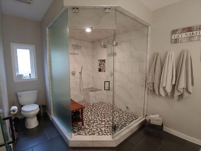 Spacious bathroom with large glass shower, pebble floor, and white marble walls. Gray walls, toilet, and towels visible.