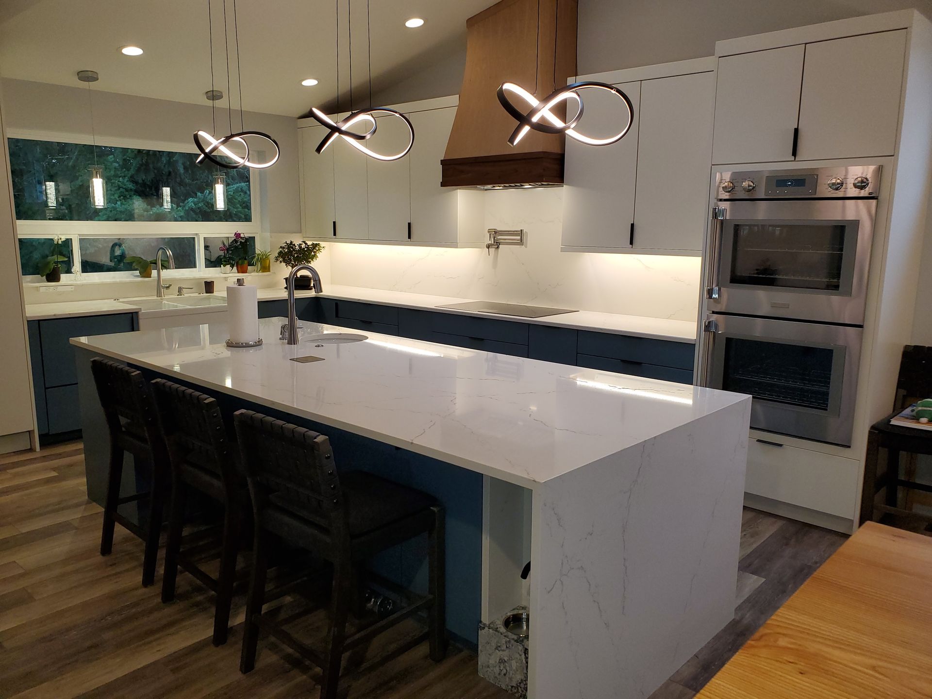 Modern kitchen with island, blue and white cabinets, wood vent hood, stainless steel appliances, and hanging lights.
