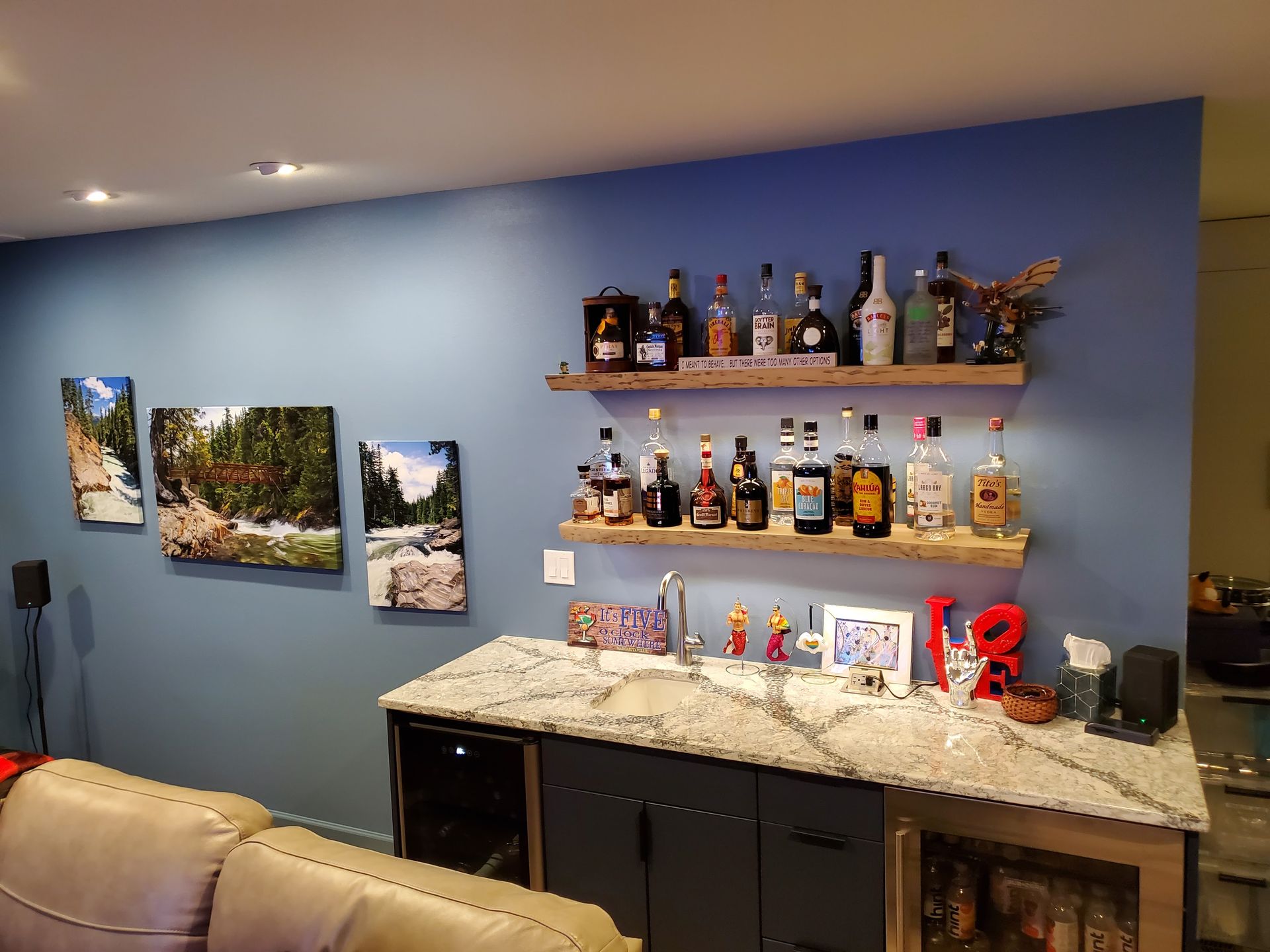 Home bar with blue wall, liquor bottles, floating shelves, granite countertop, and artwork.