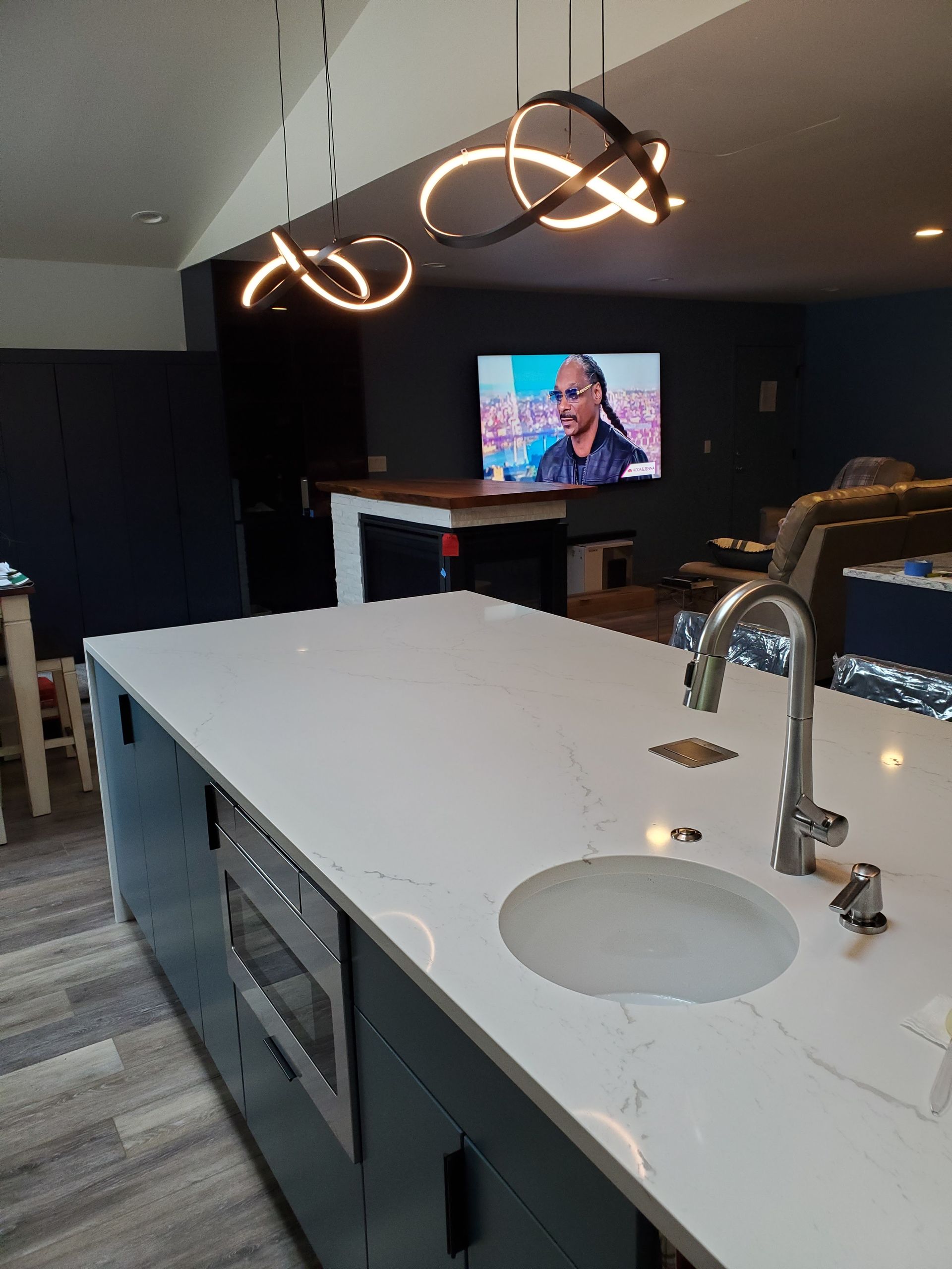 Modern kitchen with island, white countertop, sink, microwave, pendant lights, and a TV in the background.