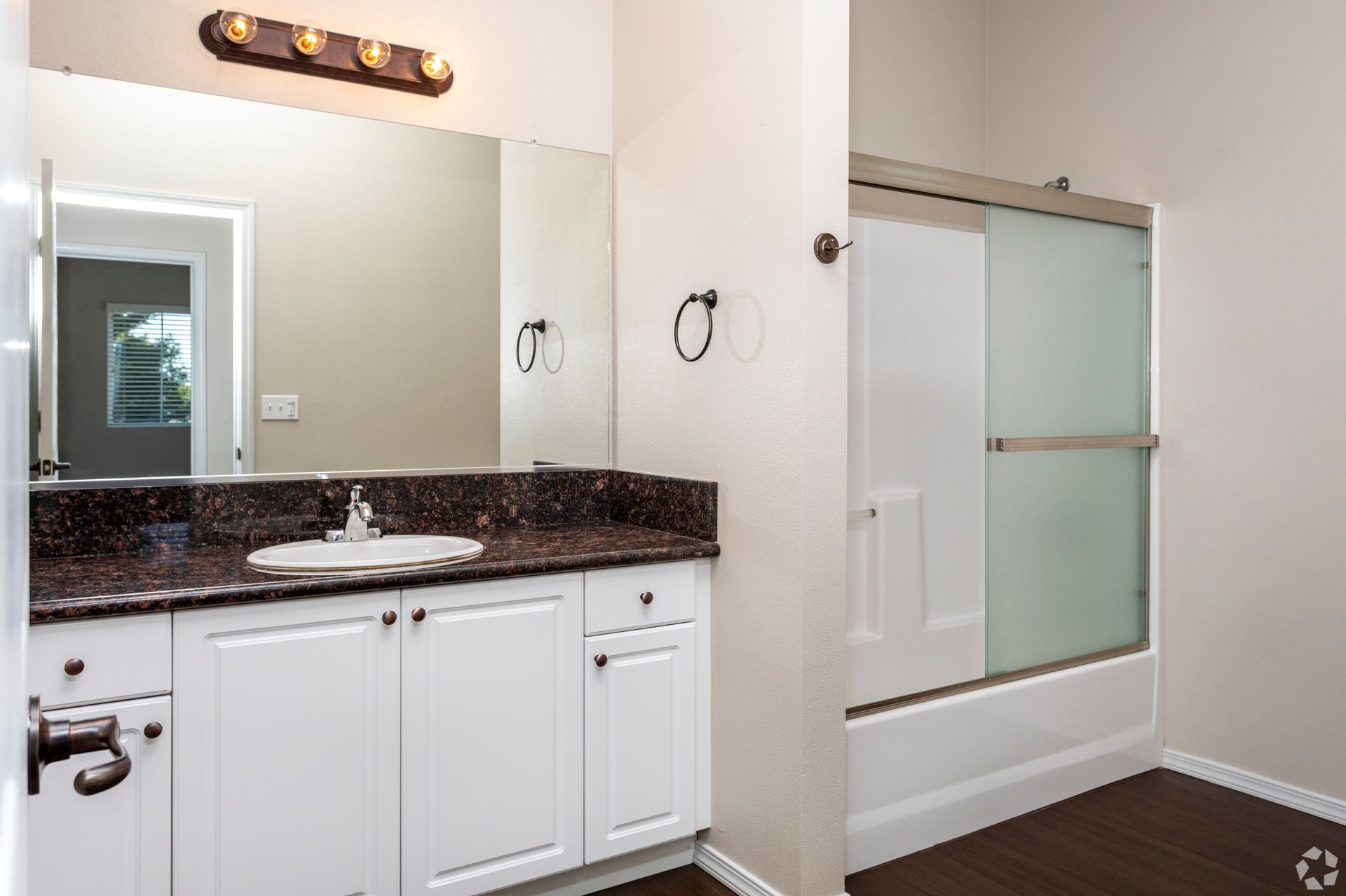 Bathroom with white vanity, brown countertop, large mirror, shower/tub combo, and brown wood floor.