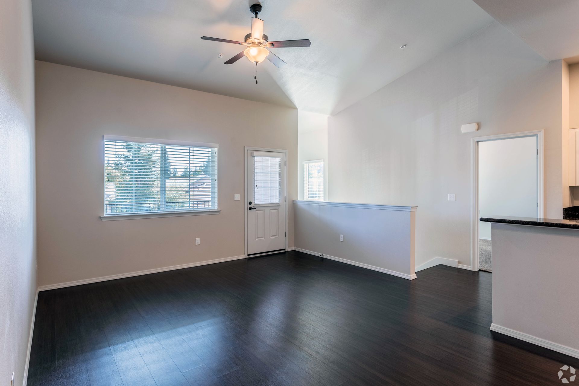 Empty living room with dark wood floor, white walls, window, door, and ceiling fan.