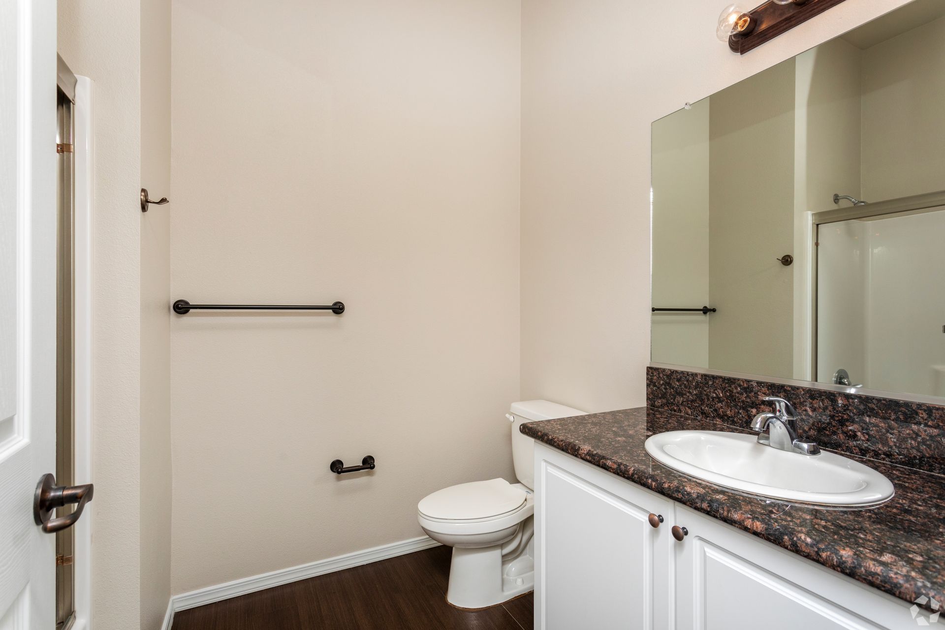 Bathroom with a toilet, sink, mirror, and towel rack. Neutral-colored walls and dark wood-look flooring.