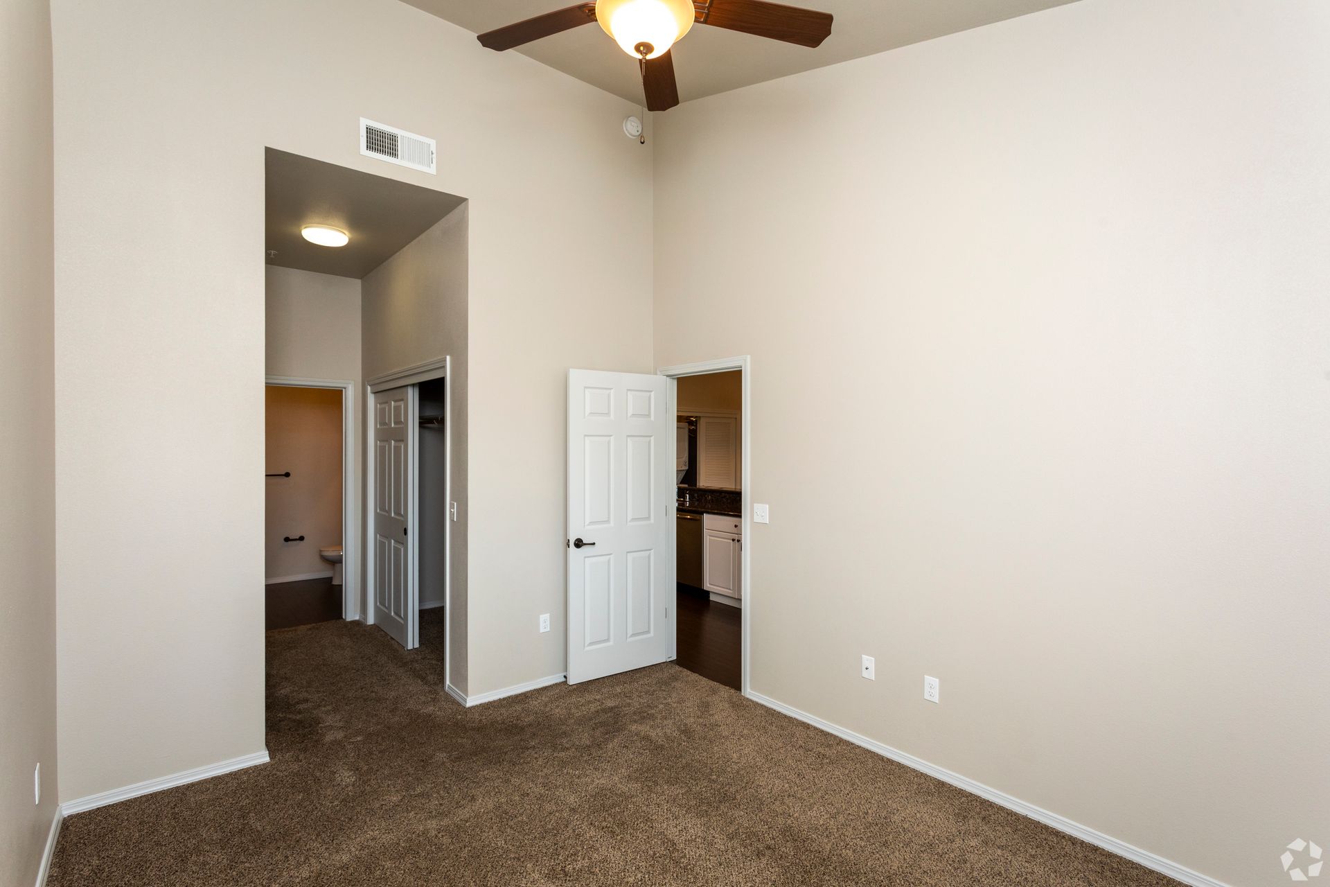 Empty bedroom with brown carpet, neutral walls, and ceiling fan; doorway to closet and kitchen.