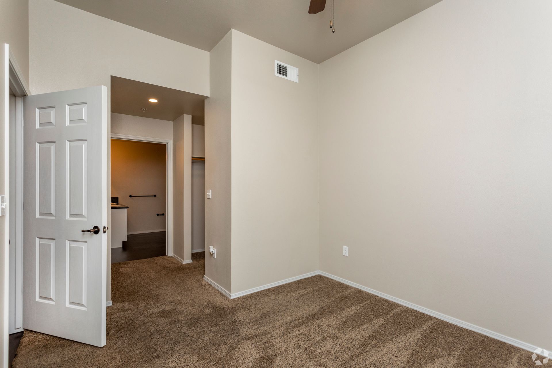 Empty room with a partially open white door leading to a hallway with a bathroom.