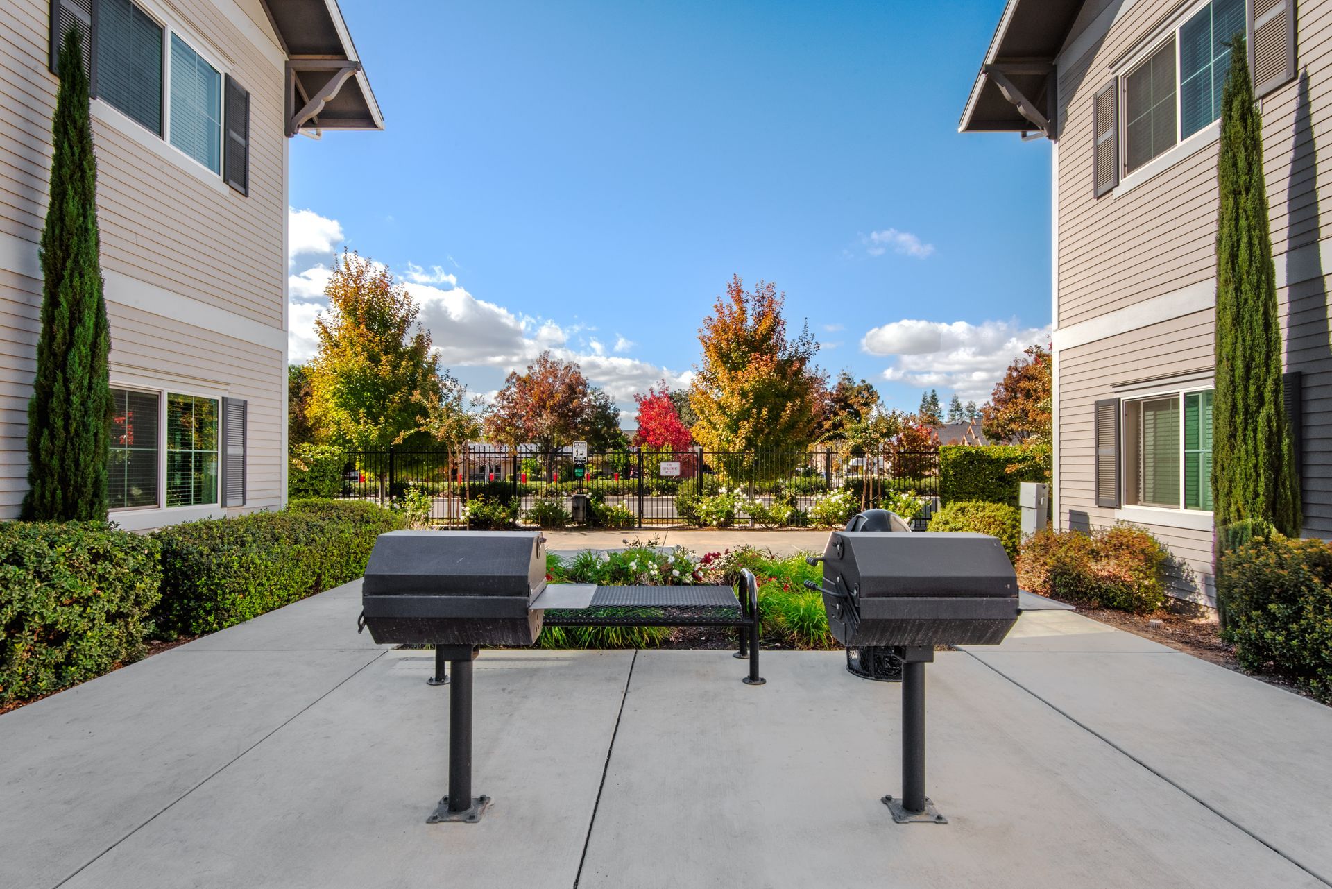 Two outdoor grills with bench, between apartment buildings under blue sky.