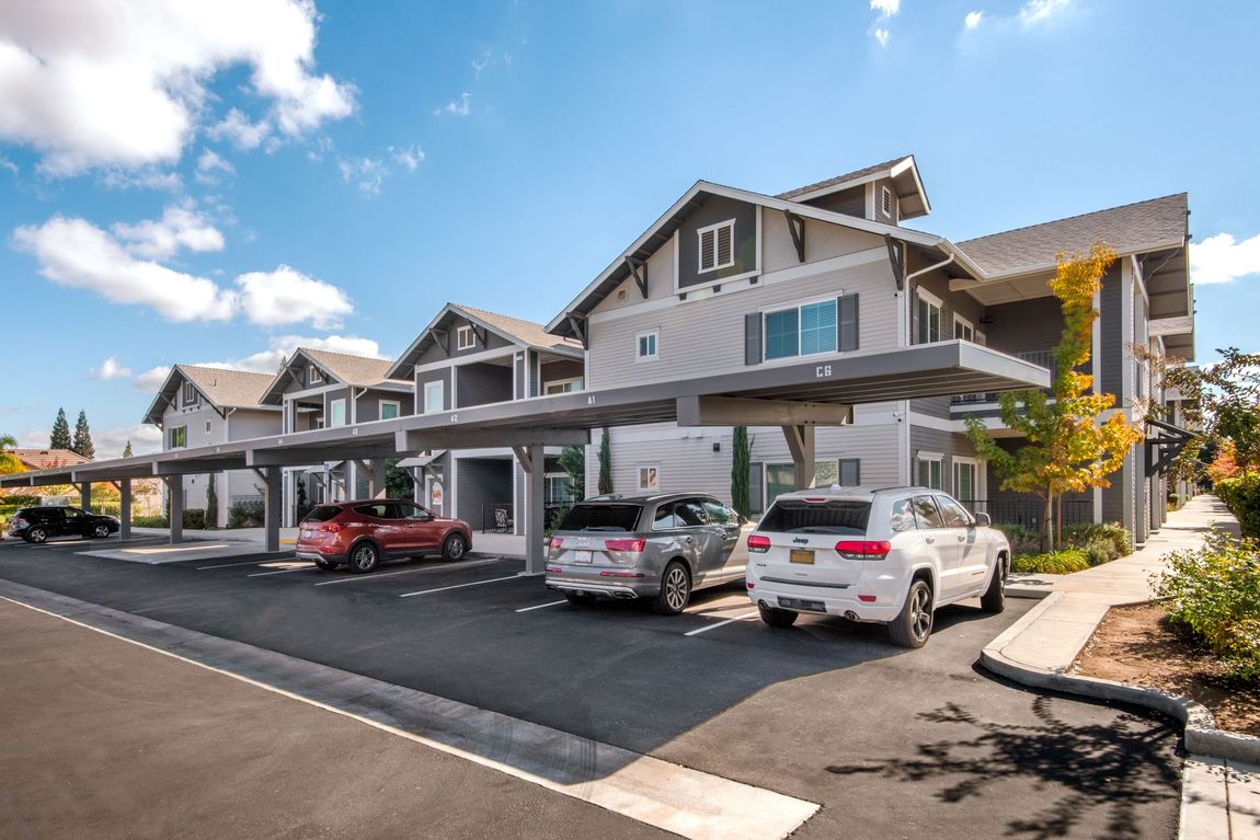Apartment complex with covered parking; cars parked under gray canopies. Sunny day, blue sky.