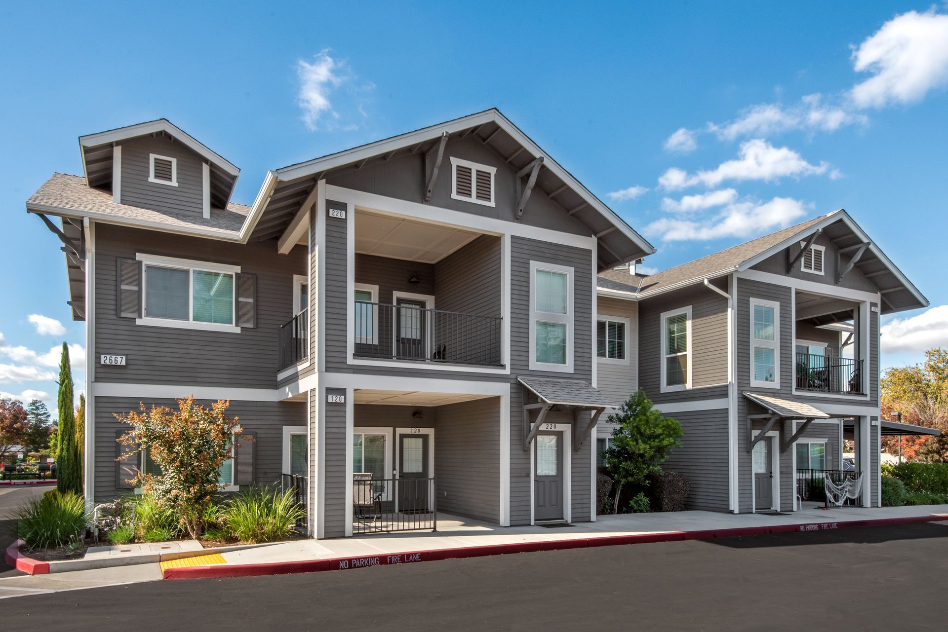 Gray two-story apartment buildings with white trim, balconies, and a blue sky.