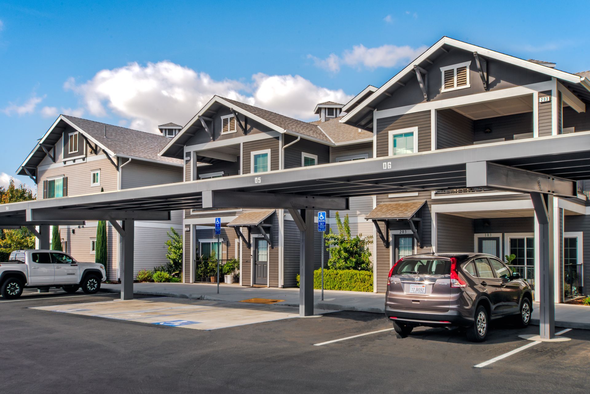 Cars parked under covered parking near grey apartment buildings. Blue sky, sunny day.