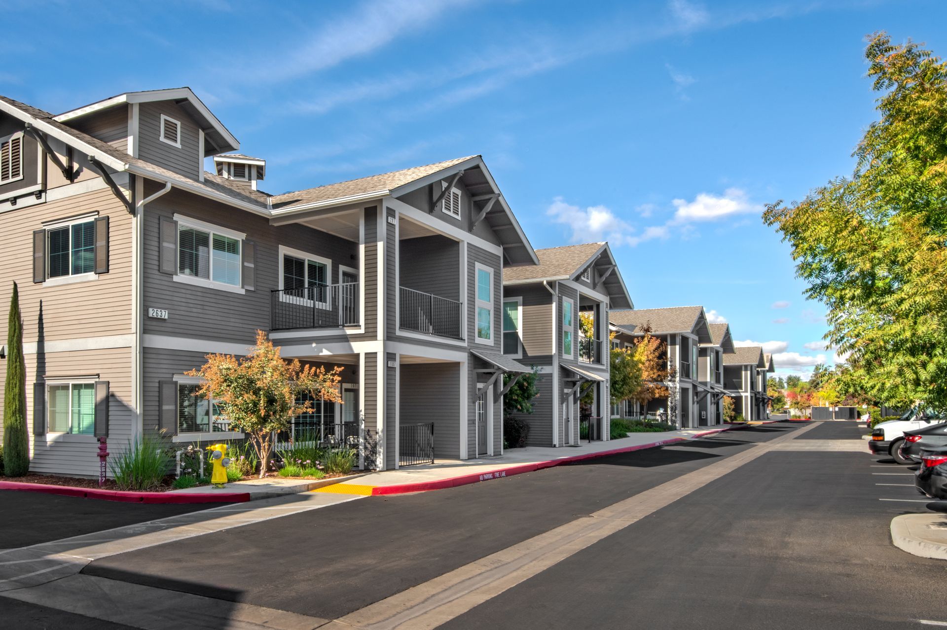 Row of multi-unit townhomes, grey siding, blue sky. Parking to the right and road in front.